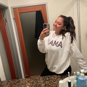 May include: A white sweatshirt with the word "MAMA" printed on the front in colorful letters. The sweatshirt is being worn by a person in a bathroom.