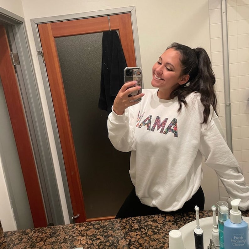 May include: A white sweatshirt with the word "MAMA" printed on the front in colorful letters. The sweatshirt is being worn by a person in a bathroom.