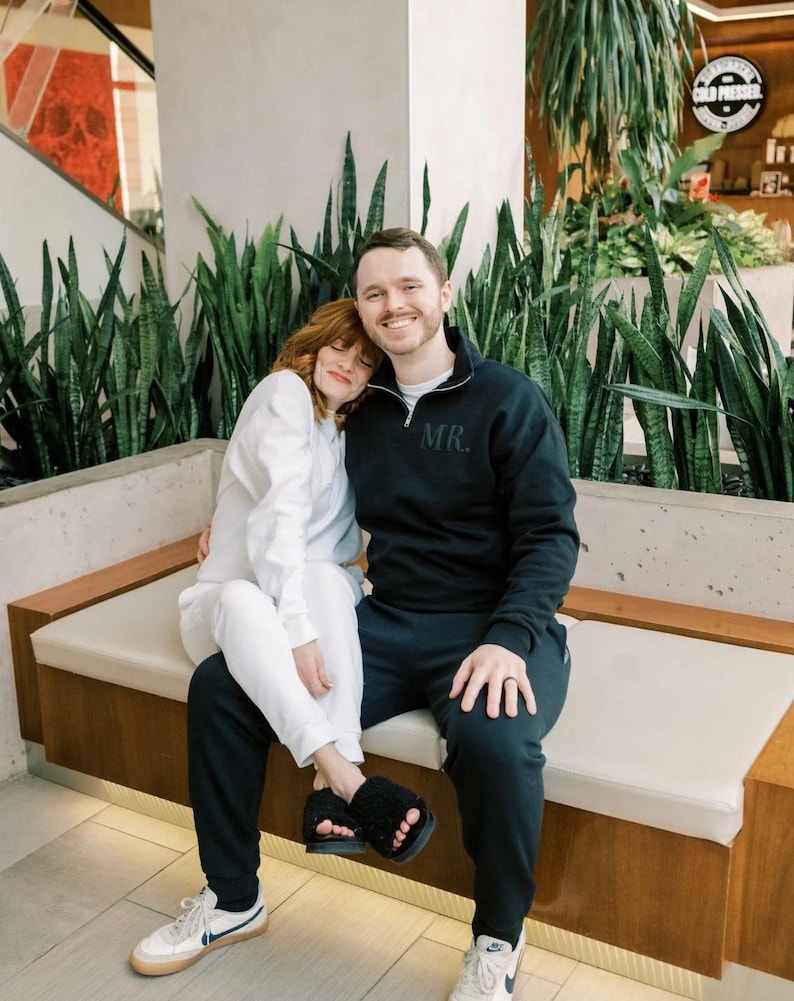 Op de afbeelding: Een stel zit op een bank met een witte achtergrond. De vrouw draagt een witte outfit en de man draagt een zwart sweatshirt met "MR." geborduurd op de borst. Ze lachen allebei.