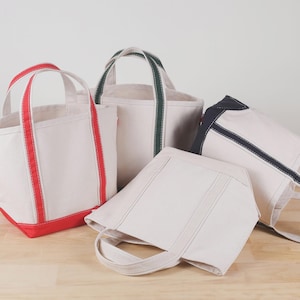 May include: Four canvas tote bags in various sizes and colors. The bags are off-white with colored accents: red, green, and navy blue. The bags have sturdy handles and are arranged on a light wood surface.