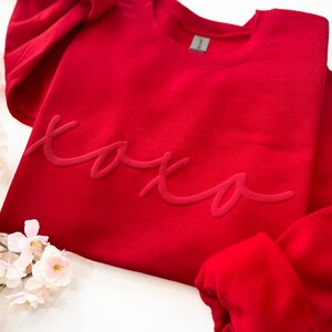 May include: A red sweatshirt with the word "xoxo" in a script font. The sweatshirt is a solid color and has a crew neck. The image also includes pink flowers.