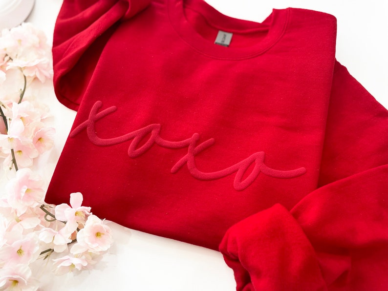 May include: A red sweatshirt with the word "xoxo" in a script font. The sweatshirt is a solid color and has a crew neck. The image also includes pink flowers.