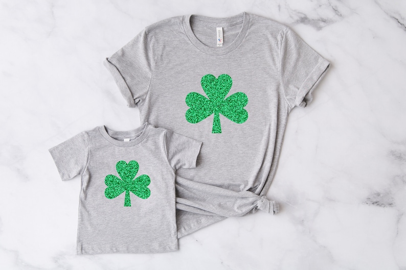 May include: Two gray t-shirts with green glitter shamrock designs. The larger shirt has a larger shamrock design and the smaller shirt has a smaller shamrock design.