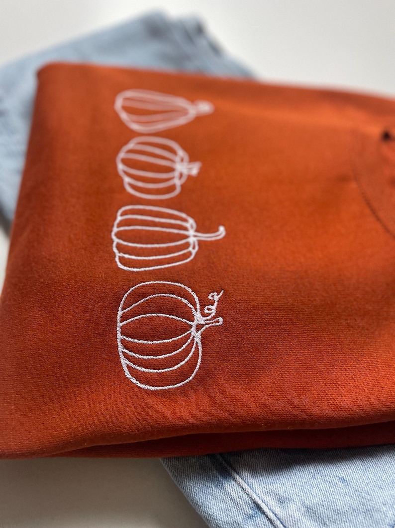 Puede incluir: Una sudadera de color &oacute;xido con un dise&ntilde;o bordado blanco de cuatro calabazas.