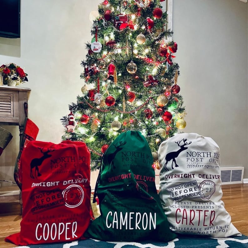 Puede incluir: Tres sacos de regalo navide&ntilde;os en rojo, verde y blanquecino, cada uno con cierre de cord&oacute;n. Los sacos presentan una silueta de reno y el texto "North Pole Post Office" y "Overnight Delivery". Los nombres "Cooper", "Cameron" y "Carter" est&aacute;n impresos en los sacos.