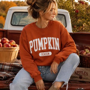 Op de afbeelding: Een roestoranje sweatshirt met lange mouwen en ronde hals, voorzien van de witte tekst PUMPKIN SEASON in een vetgedrukt, collegiaal lettertype op de borst. Het sweatshirt wordt gecombineerd met een lichtblauwe spijkerbroek en beige su&egrave;de enkellaarsjes.