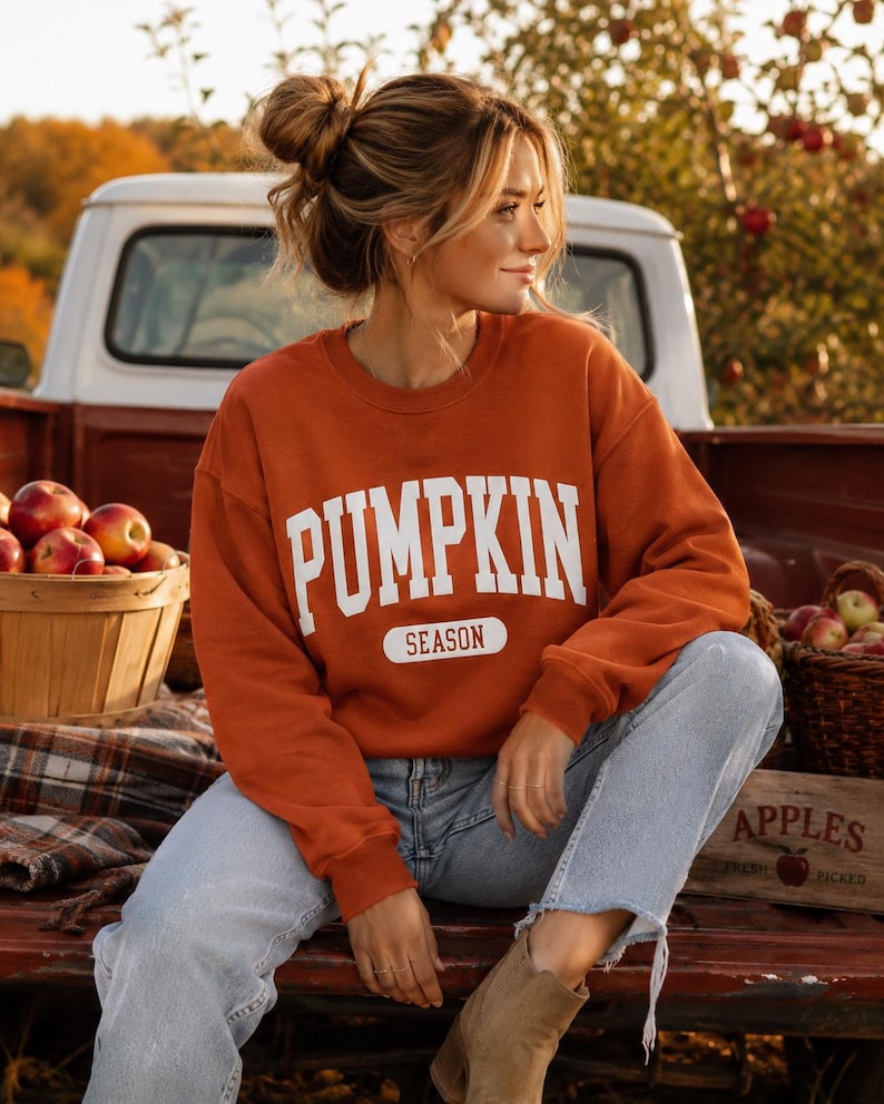 Op de afbeelding: Een roestoranje sweatshirt met lange mouwen en ronde hals, voorzien van de witte tekst PUMPKIN SEASON in een vetgedrukt, collegiaal lettertype op de borst. Het sweatshirt wordt gecombineerd met een lichtblauwe spijkerbroek en beige su&egrave;de enkellaarsjes.