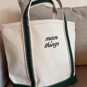 May include: A canvas tote bag with a cream-colored body and dark green accents. The bag features dark green handles and a dark green base. The words "mom things" are embroidered in black on the front of the bag.