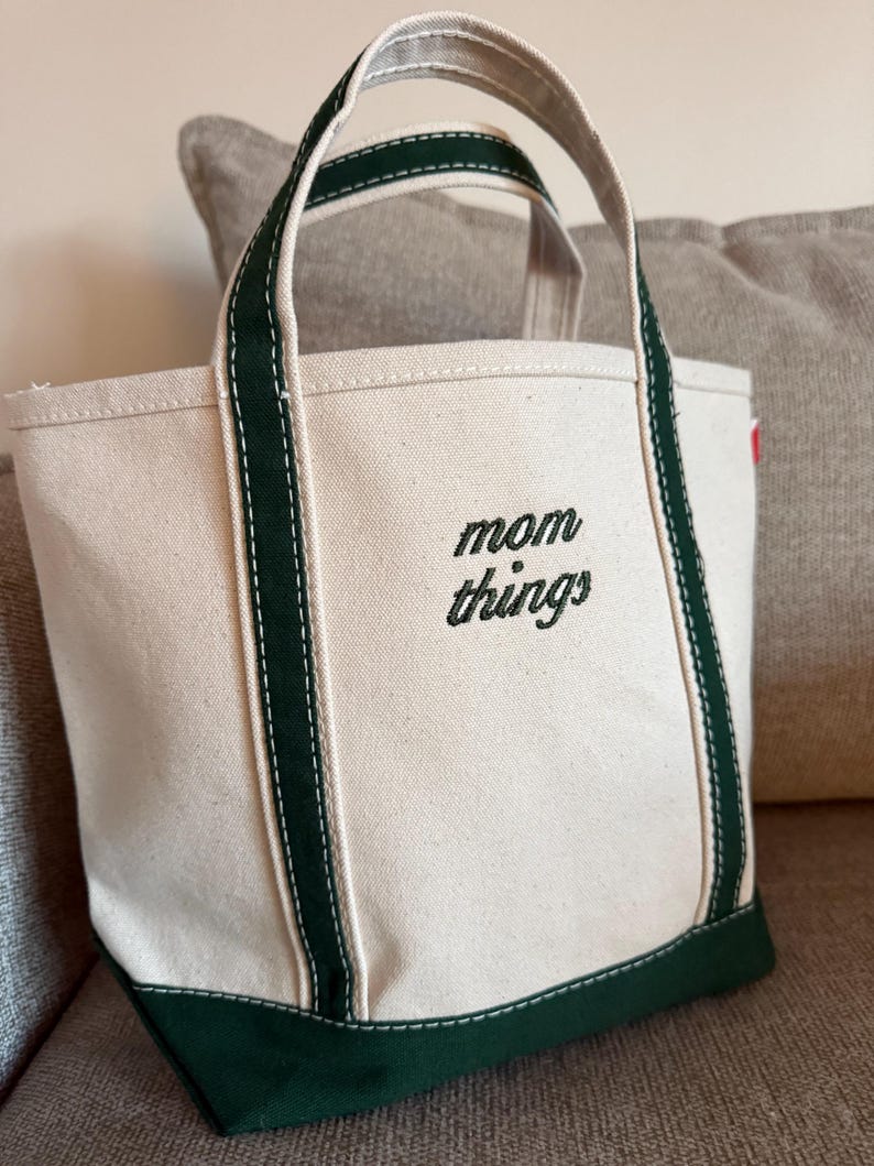 May include: A canvas tote bag with a cream-colored body and dark green accents. The bag features dark green handles and a dark green base. The words "mom things" are embroidered in black on the front of the bag.