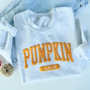 May include: A light gray sweatshirt with the words "PUMPKIN SEASON" in orange lettering. The sweatshirt is folded with the sleeves tucked in. A pair of blue jeans is behind the sweatshirt. Two decorative pumpkins are next to the sweatshirt.