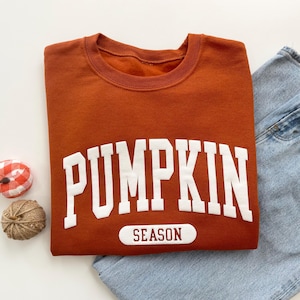 Op de afbeelding: Een roestkleurige sweater met ronde hals en de witte tekst PUMPKIN in een groot collegiaal lettertype, met het woord SEASON in donkerrood gedrukt op een witte rechthoekige patch eronder. De sweater wordt gecombineerd met een lichtblauwe jeans.