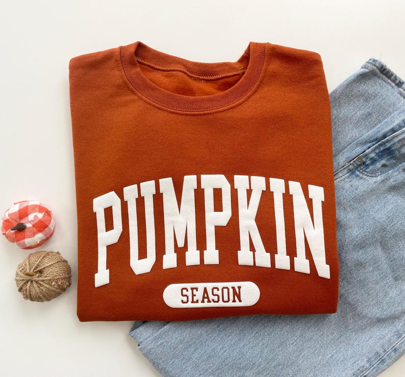 Op de afbeelding: Een roestkleurige sweater met ronde hals en de witte tekst PUMPKIN in een groot collegiaal lettertype, met het woord SEASON in donkerrood gedrukt op een witte rechthoekige patch eronder. De sweater wordt gecombineerd met een lichtblauwe jeans.