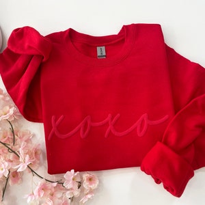 May include: A vibrant red sweatshirt with the word "xoxo" in a tone-on-tone script. The long-sleeved top is displayed with white sneakers and pink flowers, creating a casual and romantic aesthetic. The sweatshirt is made of a soft material.