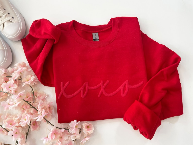 May include: A vibrant red sweatshirt with the word "xoxo" in a tone-on-tone script. The long-sleeved top is displayed with white sneakers and pink flowers, creating a casual and romantic aesthetic. The sweatshirt is made of a soft material.