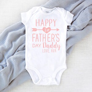 Peut inclure: Combinaison blanche pour b&eacute;b&eacute; avec un texte rose qui dit "Happy 1st Father's Day Daddy Love, Ava". Le texte est entour&eacute; d'un c&oelig;ur rose et d'une fl&egrave;che.