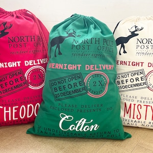 Puede incluir: Tres sacos navide&ntilde;os con cord&oacute;n en rojo, verde y crema. Cada saco presenta una silueta de reno, el texto "North Pole Post Office" y "Overnight Delivery". Los sacos est&aacute;n personalizados con nombres.
