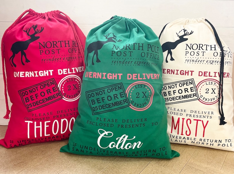 Puede incluir: Tres sacos navide&ntilde;os con cord&oacute;n en rojo, verde y crema. Cada saco presenta una silueta de reno, el texto "North Pole Post Office" y "Overnight Delivery". Los sacos est&aacute;n personalizados con nombres.