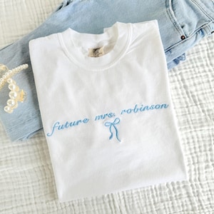 White Comfort Colors t-shirt with light blue embroidered text reading “future mrs. robinson” and a delicate bow, folded on a white textured blanket next to light wash jeans and pearl hair clips – perfect custom bridal shirt or engagement gift.