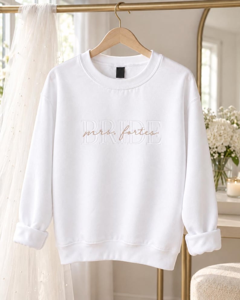 May include: A white long-sleeve crewneck sweatshirt featuring the word BRIDE embroidered in large white block letters across the chest, with the name mrs. fortes embroidered in a smaller, cursive gold script layered over the center.