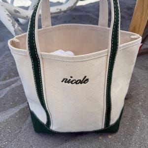 May include: Cream-colored canvas tote bag with dark green accents on the base and handles. The word "nicole" is embroidered in black script on the front of the bag. The bag is sitting on a sandy surface.