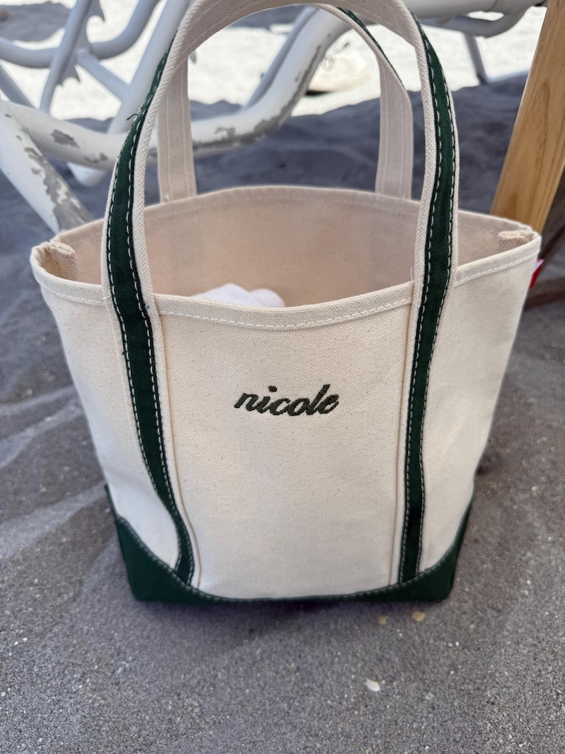 May include: Cream-colored canvas tote bag with dark green accents on the base and handles. The word "nicole" is embroidered in black script on the front of the bag. The bag is sitting on a sandy surface.