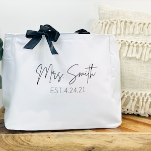 K&ouml;nnte beinhalten: Wei&szlig;e Canvas-Tragetasche mit schwarzen Schleifenb&auml;ndern als Henkeln und dem in Schwarz gedruckten Text "Mrs. Smith EST. 4.24.21".