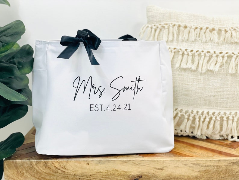 K&ouml;nnte beinhalten: Wei&szlig;e Canvas-Tragetasche mit schwarzen Schleifenb&auml;ndern als Henkeln und dem in Schwarz gedruckten Text "Mrs. Smith EST. 4.24.21".