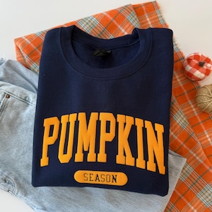 Op de afbeelding: Een marineblauw sweatshirt met ronde hals waarop het woord PUMPKIN in grote, dikke, oranje hoofdletters in een boog over de borst staat, met het woord SEASON in kleinere zwarte letters in een oranje rechthoekig label eronder.