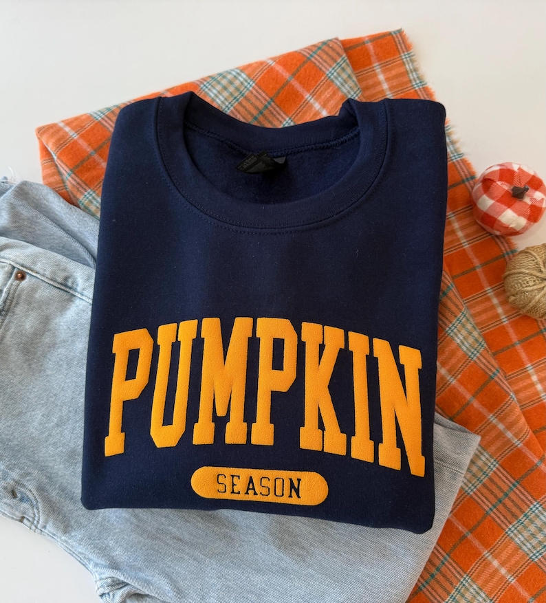 Op de afbeelding: Een marineblauw sweatshirt met ronde hals waarop het woord PUMPKIN in grote, dikke, oranje hoofdletters in een boog over de borst staat, met het woord SEASON in kleinere zwarte letters in een oranje rechthoekig label eronder.