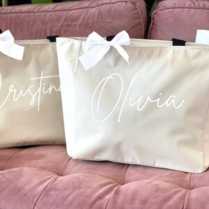 May include: Three beige canvas tote bags with white ribbon bows and black handles. The bags are personalized with the names "Cristina", "Olivia", and "A" in white script.
