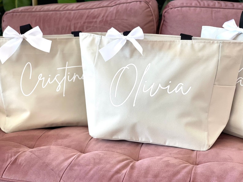 May include: Three beige canvas tote bags with white ribbon bows and black handles. The bags are personalized with the names "Cristina", "Olivia", and "A" in white script.