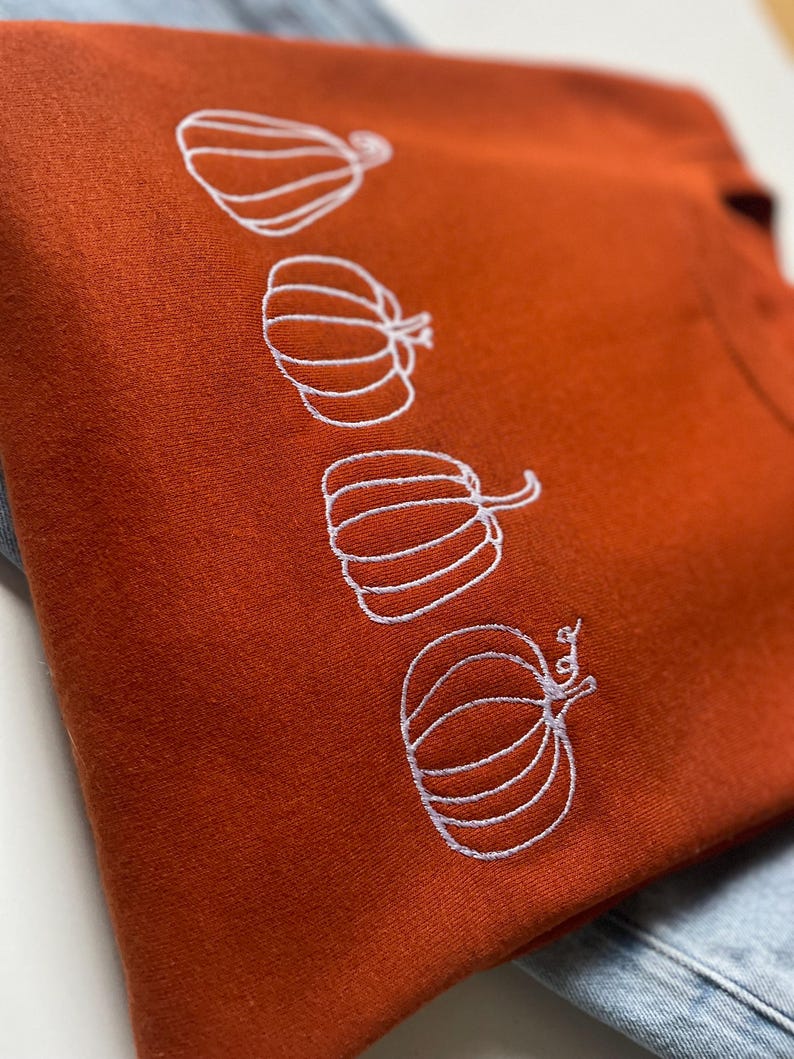 Puede incluir: Una sudadera de color &oacute;xido con un dise&ntilde;o bordado blanco de cuatro calabazas.