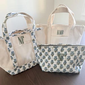 May include: Two canvas tote bags and one quilted zippered pouch. One tote features a blue and green floral trim and the monogram NF. The second tote is plain off-white canvas with the monogram KMR. The pouch has a blue and green floral pattern and the monogram NT.