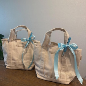 May include: Two off-white canvas tote bags with two top handles each. The left bag features the embroidered name "Heeroni" and the right bag features "Wuch". Both bags are decorated with a light blue satin ribbon bow and a white hair claw clip.