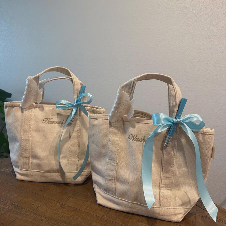 May include: Two off-white canvas tote bags with two top handles each. The left bag features the embroidered name "Heeroni" and the right bag features "Wuch". Both bags are decorated with a light blue satin ribbon bow and a white hair claw clip.