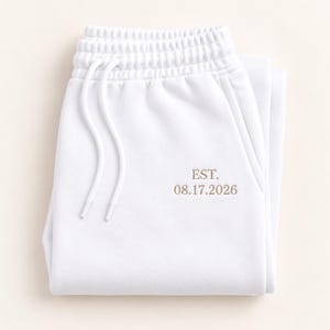May include: White sweatpants featuring an elastic waistband with matching white drawstring cords. The front of the garment is embroidered with the text EST. 08.17.2026 in a light brown thread color, positioned near the right side pocket area.