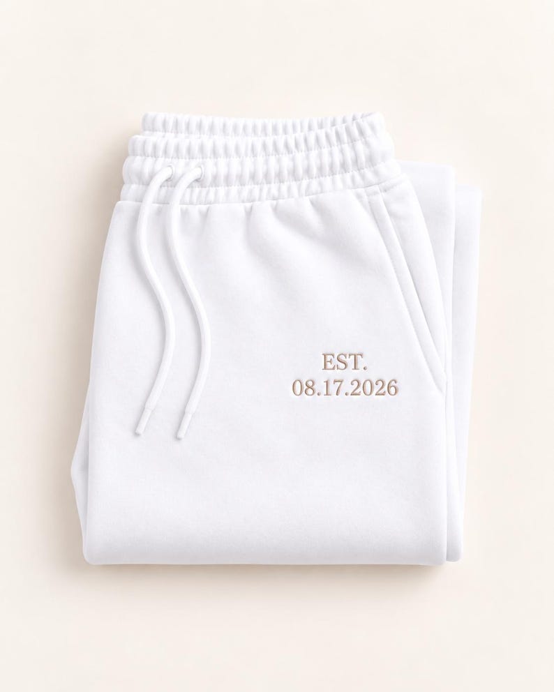 May include: White sweatpants featuring an elastic waistband with matching white drawstring cords. The front of the garment is embroidered with the text EST. 08.17.2026 in a light brown thread color, positioned near the right side pocket area.