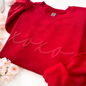 May include: A red sweatshirt with the word "xoxo" in a tonal, raised script. The sweatshirt is folded, with a portion of the sleeve visible. White sneakers and pink flowers are in the background.