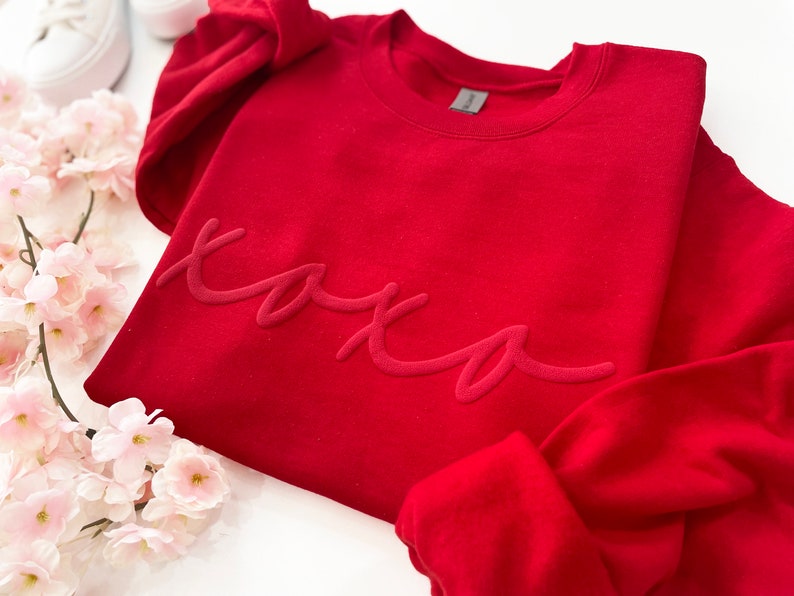 May include: A red sweatshirt with the word "xoxo" in a tonal, raised script. The sweatshirt is folded, with a portion of the sleeve visible. White sneakers and pink flowers are in the background.
