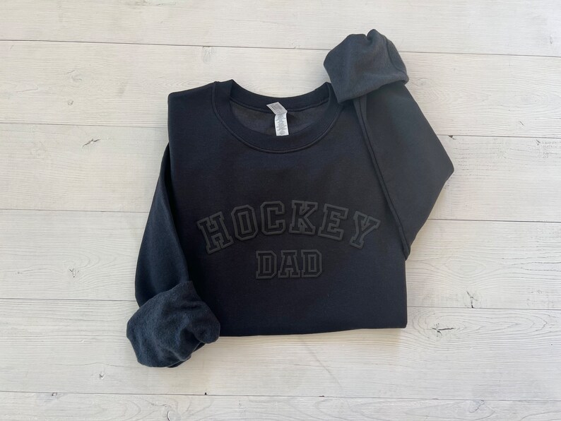 K&ouml;nnte beinhalten: Schwarzer Pullover mit dem Text "HOCKEY DAD" in wei&szlig;er Schrift mit schwarzem Umriss.
