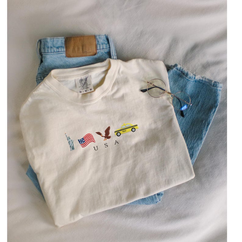 Puede incluir: Camiseta color crema con bordados tem&aacute;ticos de EE. UU., incluyendo la Estatua de la Libertad, una bandera estadounidense, un &aacute;guila y un taxi amarillo. La camiseta se combina con vaqueros azules y gafas de montura redonda.