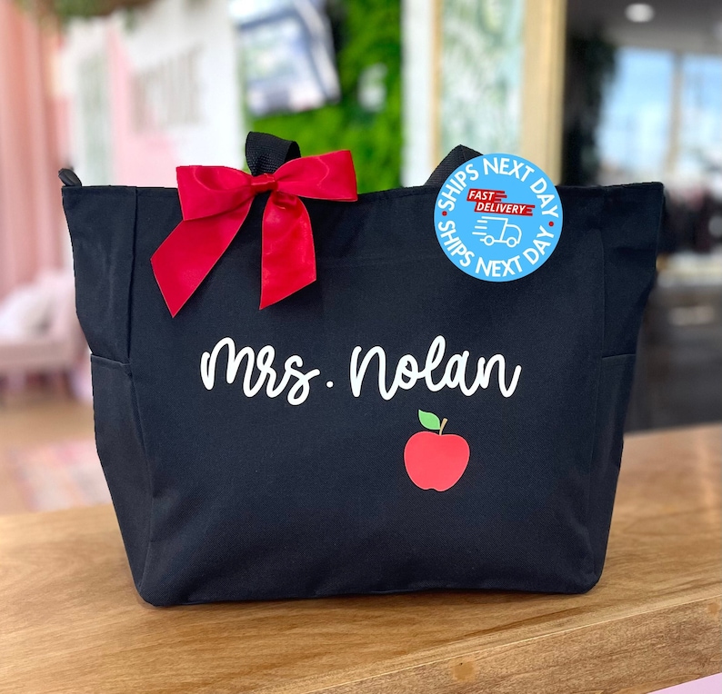 May include: Black tote bag with a red bow, a red apple graphic, and the text "Mrs. Nolan". The bag has a blue circle sticker that says "Ships Next Day Fast Delivery Ships Next Day".