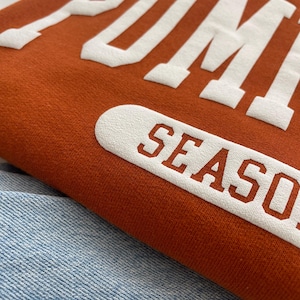 Op de afbeelding: Een roestoranje sweatshirt met grote, witte, verhoogde blokletters die PUMPKIN SEASON spellen. De stof heeft een zachte, getextureerde uitstraling en het kledingstuk ligt op een lichtblauwe spijkerbroek met een zichtbaar weefpatroon.