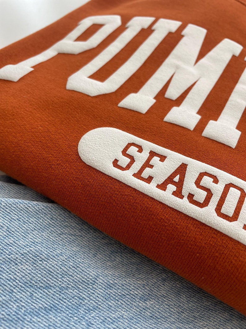 Op de afbeelding: Een roestoranje sweatshirt met grote, witte, verhoogde blokletters die PUMPKIN SEASON spellen. De stof heeft een zachte, getextureerde uitstraling en het kledingstuk ligt op een lichtblauwe spijkerbroek met een zichtbaar weefpatroon.