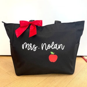 May include: Black tote bag with a red bow and the text "Mrs. Nolan" in white. A red apple with a green stem is below the text.