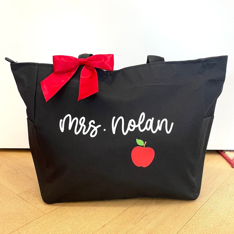 May include: Black tote bag with a red bow and the text "Mrs. Nolan" in white. A red apple with a green stem is below the text.
