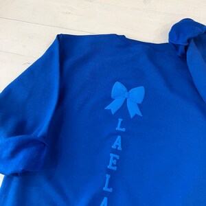 May include: Royal blue sweatshirt with a blue bow and the name "LAELLA" spelled vertically down the back.