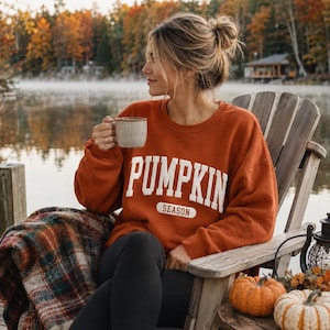 Op de afbeelding: Een roestoranje sweater met ronde hals en de witte tekst PUMPKIN SEASON op de borst in een vetgedrukt, collegiaal lettertype. Het kledingstuk heeft lange mouwen en een relaxte pasvorm, gemaakt van een effen gekleurde stof.