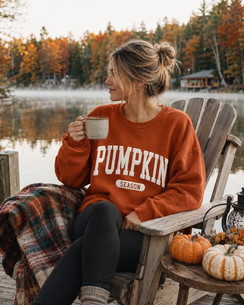 Op de afbeelding: Een roestoranje sweater met ronde hals en de witte tekst PUMPKIN SEASON op de borst in een vetgedrukt, collegiaal lettertype. Het kledingstuk heeft lange mouwen en een relaxte pasvorm, gemaakt van een effen gekleurde stof.