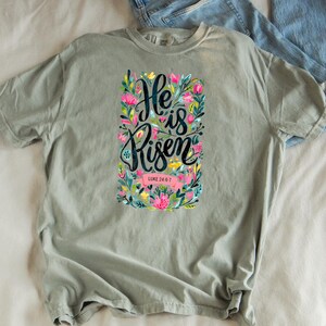 May include: A light green t-shirt with a floral design and the text "He is Risen" with the scripture reference "Luke 24:6-7".